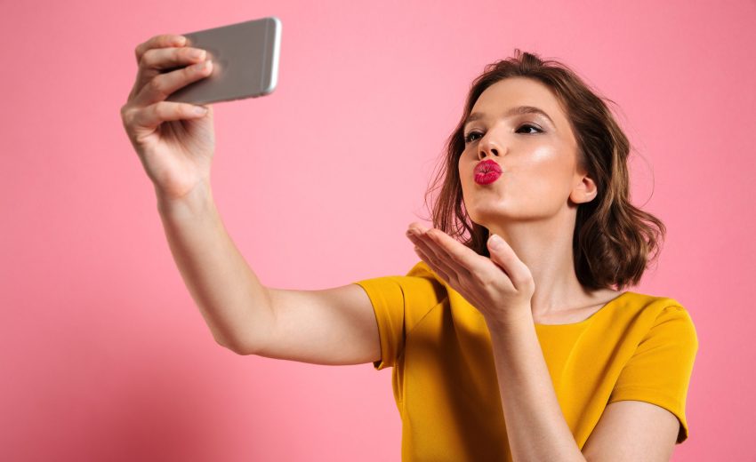 Close-up shot of young attractive woman with bright makeup sending air kiss while taking selfie on mobile phone, isolated over pink background